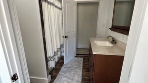 Un couloir avec un plancher en bois, un tapis blanc, une salle de bain avec un lavabo et un miroir, et une porte donnant sur une autre pièce.
