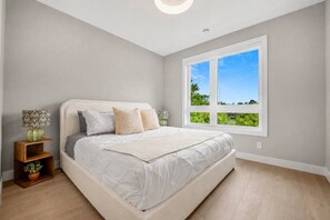 Et soveværelse med et stort vindue, en seng med hvide sengetøj, et natbord med en lampe og en lille plante.