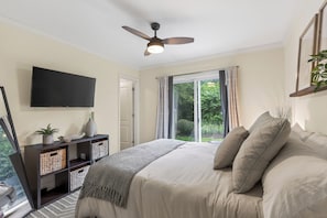Une chambre à coucher avec un lit, un ventilateur de plafond, une télévision, une fenêtre avec des rideaux et une plante.