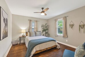 Une chambre à coucher avec un lit, un ventilateur de plafond, une fenêtre avec des rideaux, une lampe de chevet et une plante.