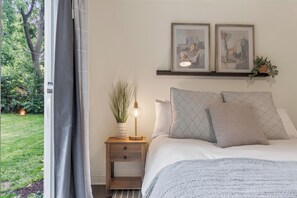 Un dormitorio con cama, mesita de noche, lámpara, planta y dos cuadros en la pared.