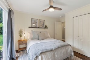Une chambre à coucher comprenant un lit, une table de chevet, un ventilateur de plafond, une fenêtre avec des rideaux et une porte.