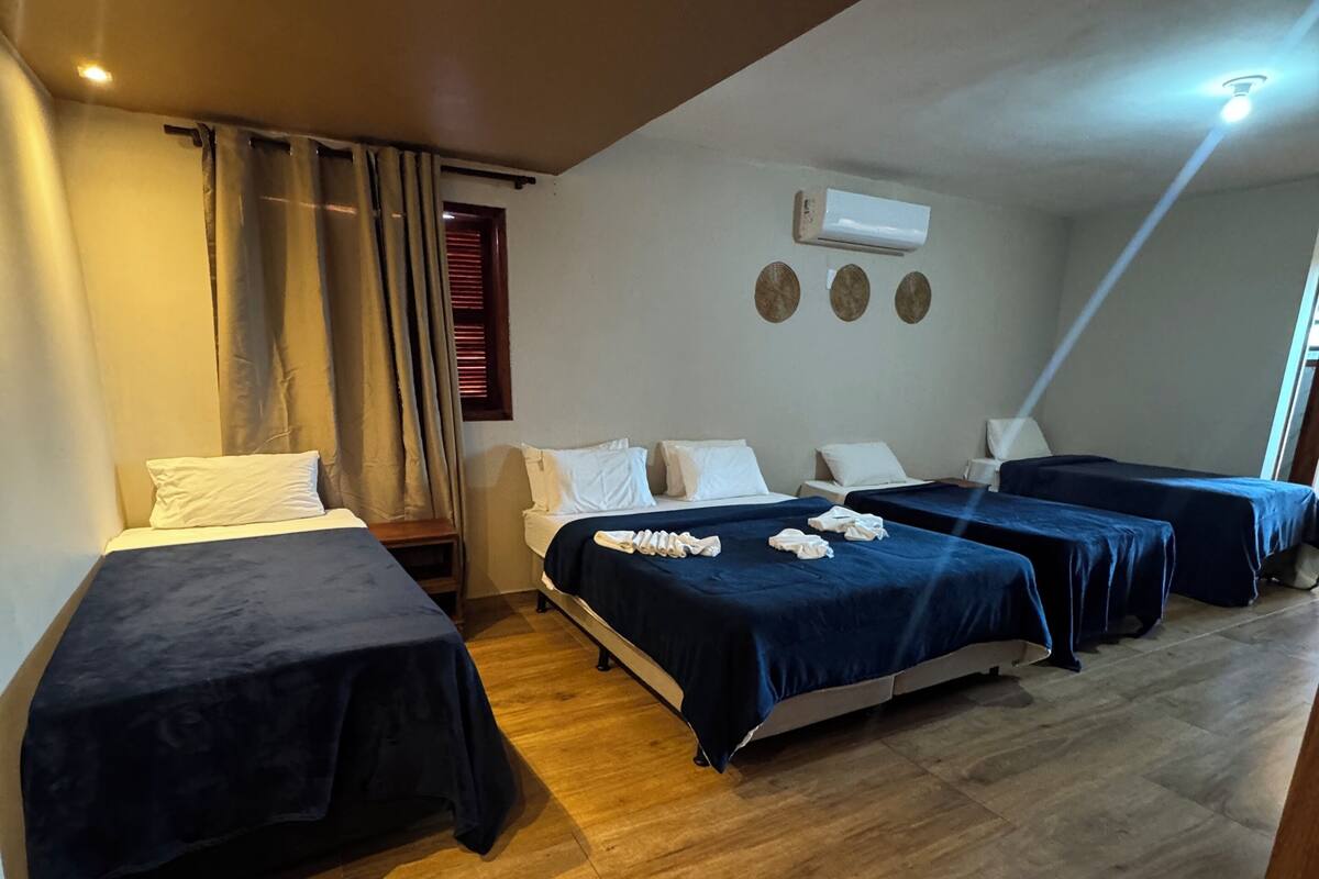 Una habitación de hotel con tres camas, suelo de madera, unidad de aire acondicionado y una ventana con cortinas.