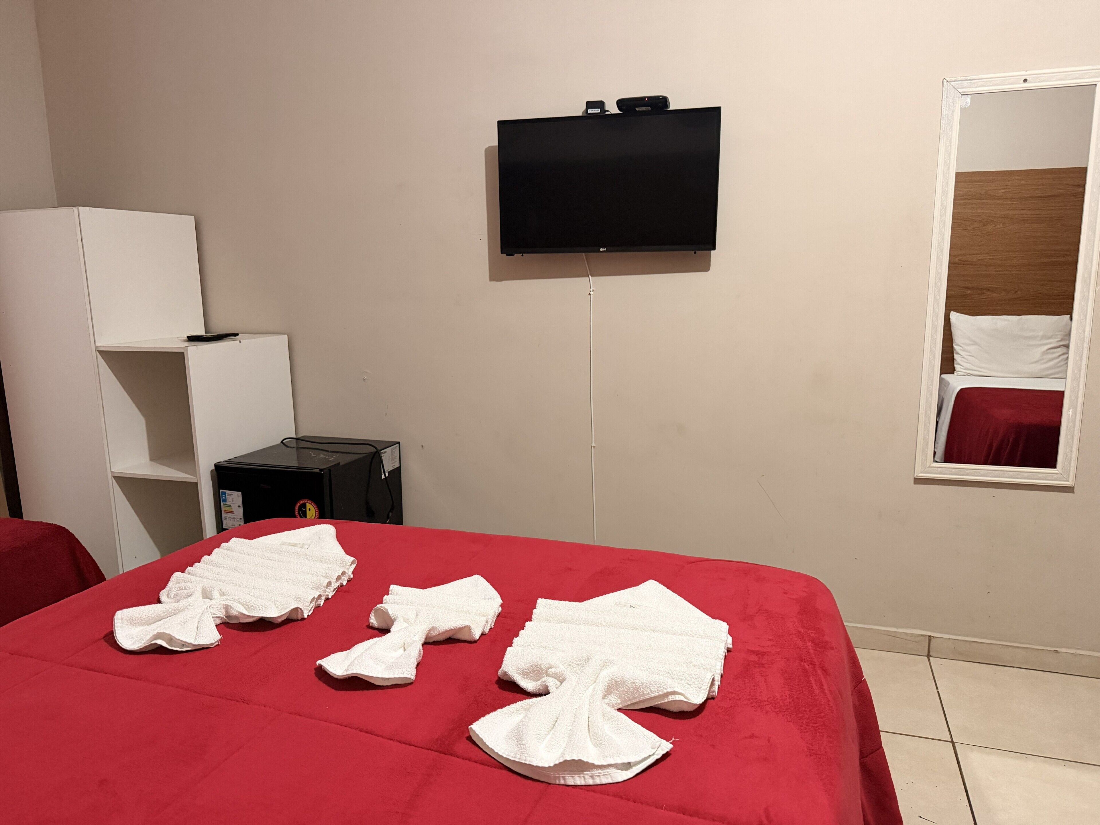 A hotel room with a bed, a TV mounted on the wall, a mirror, and a small desk with a lamp.