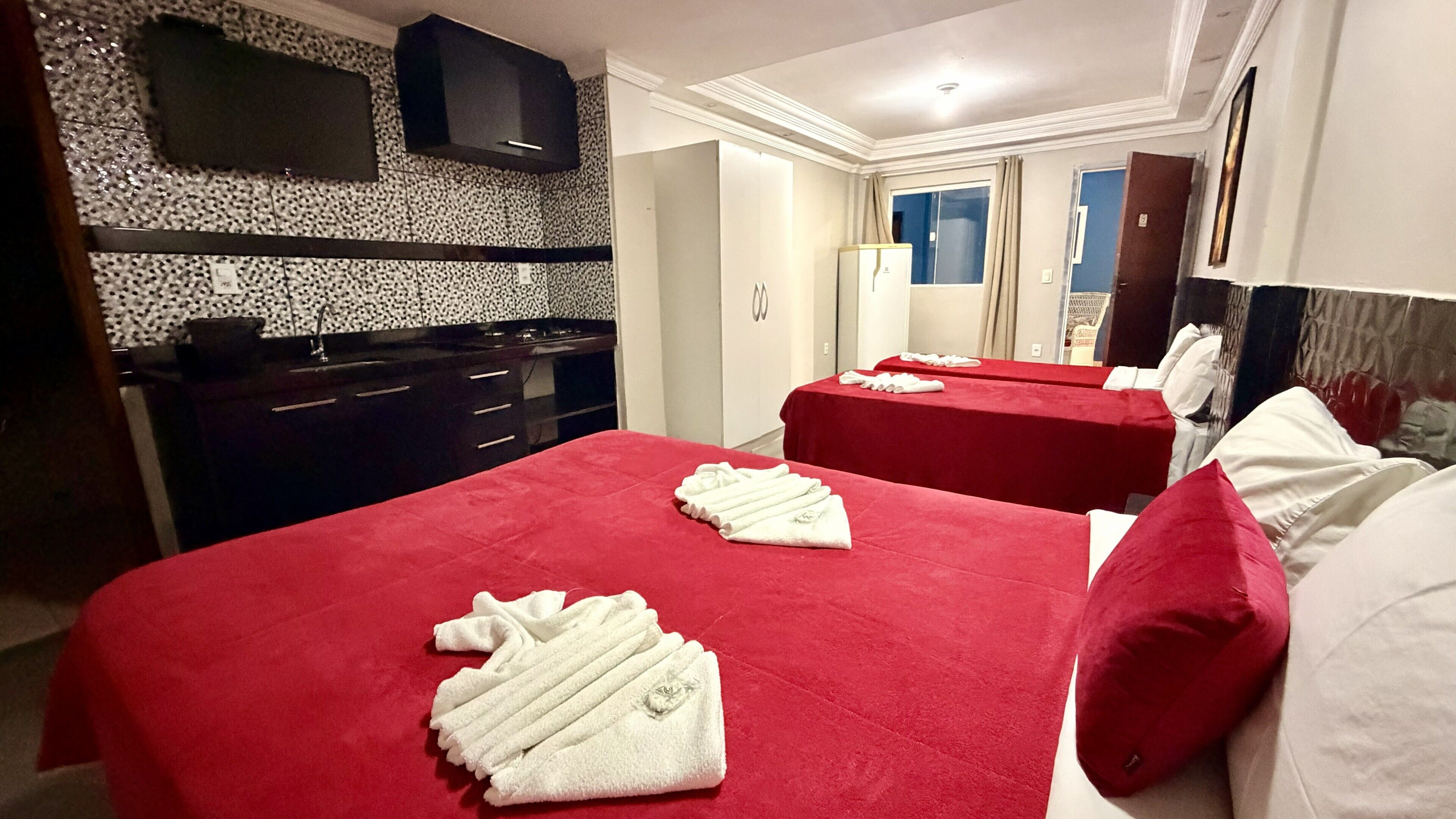 Quarto de hotel com duas camas, roupa de cama vermelha, área de cozinha com bancada preta e uma televisão na parede.