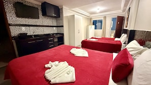Habitación de hotel con dos camas, ropa de cama roja, una cocina con encimera negra y un televisor montado.