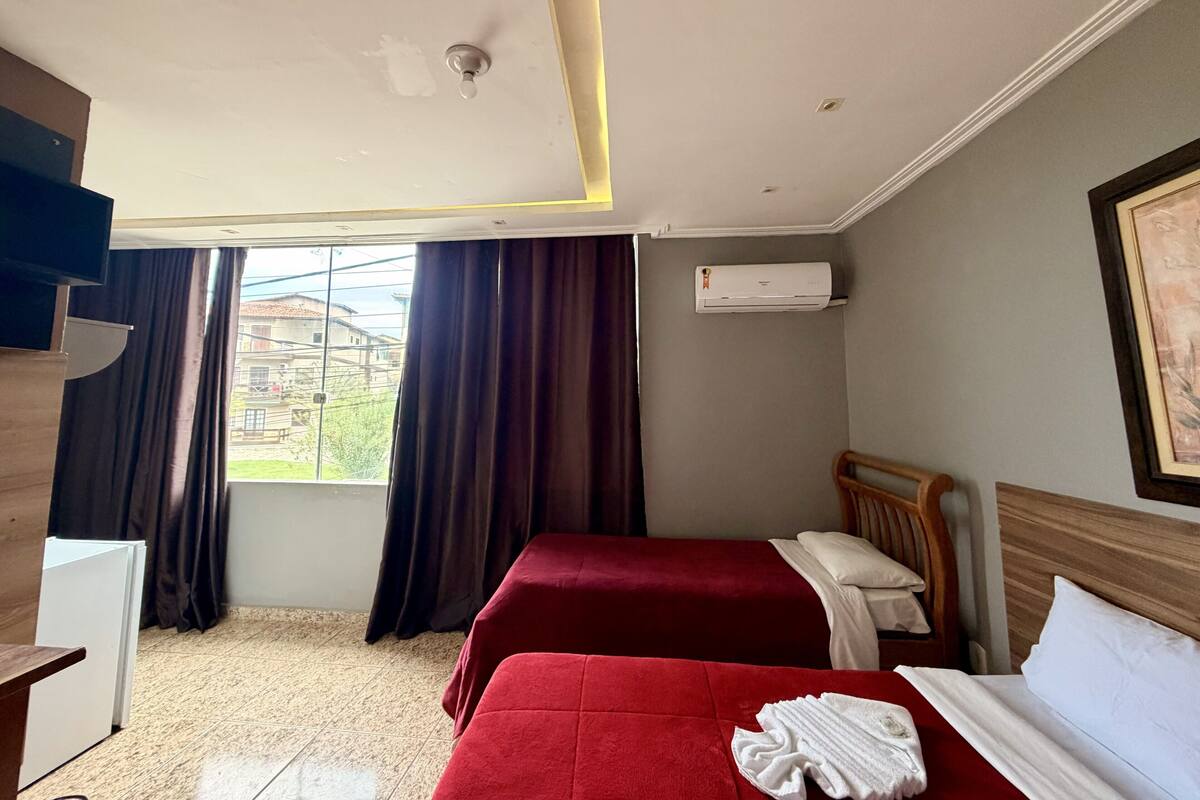 Habitación de hotel con dos camas, una ventana con cortinas, un equipo de aire acondicionado y un cuadro en la pared.