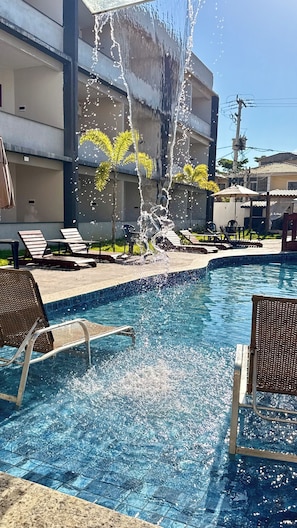 A swimming pool with a water fountain, lounge chairs, and a building in the background. - Maresia Parque Hotel (Rio das Ostras)