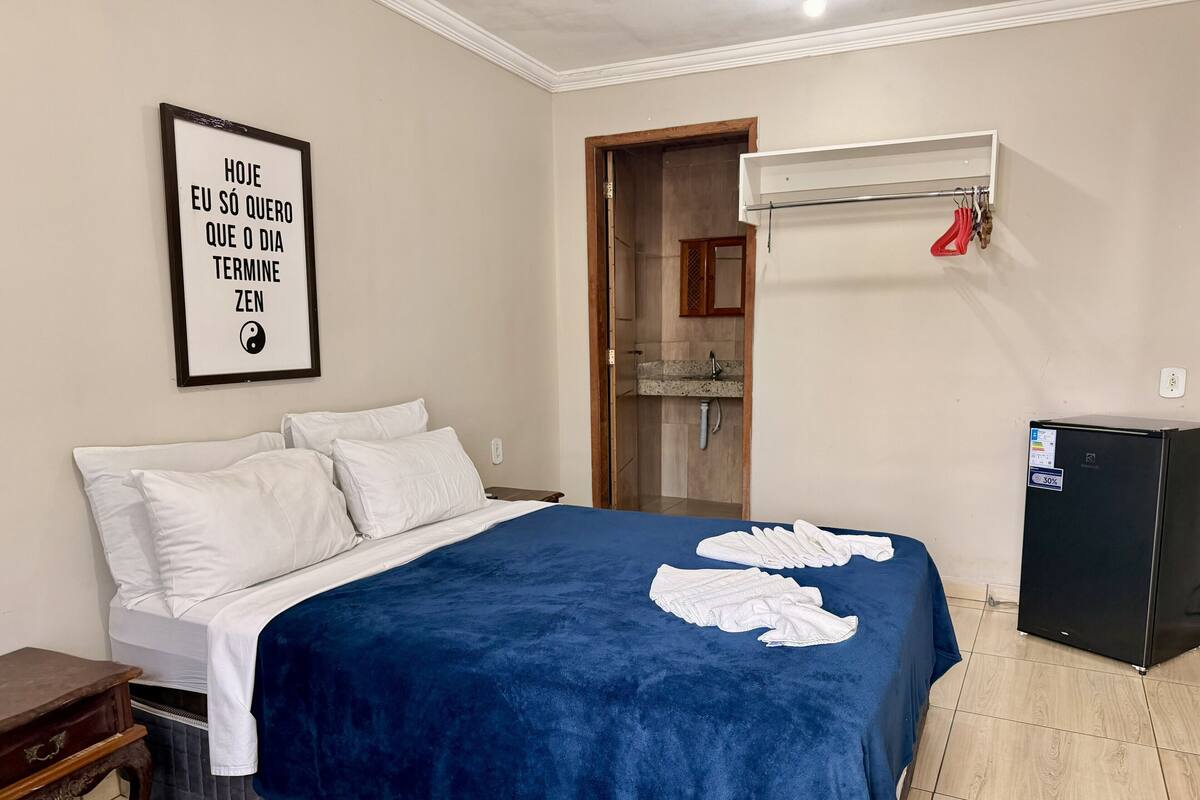Un dormitorio con cama, mesita de noche, aire acondicionado instalado en la pared, un refrigerador pequeño y un baño visible a través de una puerta abierta.