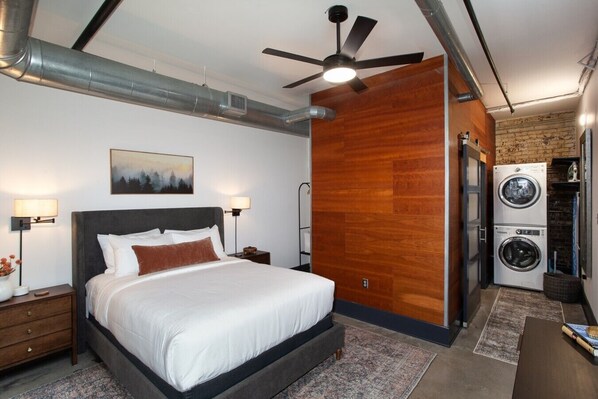 A bedroom with a bed, nightstands, a ceiling fan, a painting, and a laundry area with a washing machine. - Stylish Loft on South Gay | Downtown Knoxville (Knoxville)