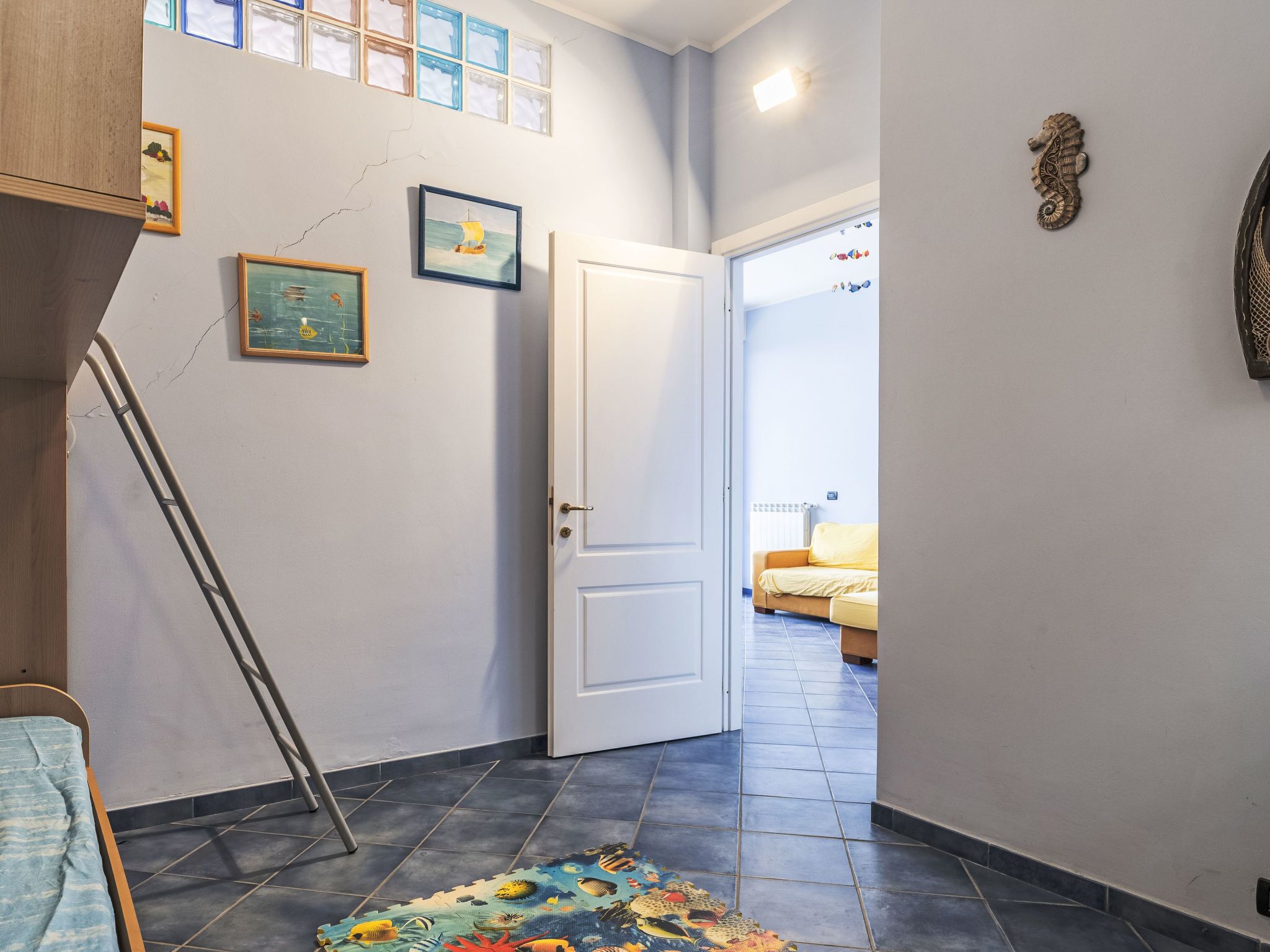 Un corridoio con una porta bianca, pavimento piastrellato, un letto con coperta gialla e una decorazione a cavalluccio marino sul muro.