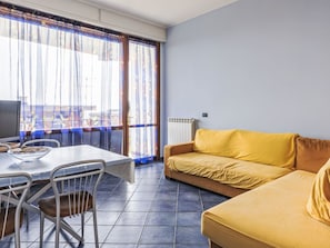 Ein Wohnzimmer mit einer Couch, einem Esstisch mit Stühlen, einem Fernseher und Blick durch ein Fenster mit Vorhängen.