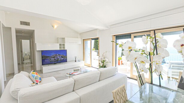 Ein modernes Wohnzimmer mit einer Couch, einem Glastisch, einem Flachbildfernseher und Blick ins Freie.