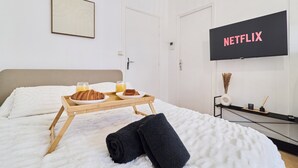 Una habitación de hotel con una cama, una bandeja con croissants y jugo, una pantalla de televisión mostrando 'NETFLIX' y un cuadro en la pared.