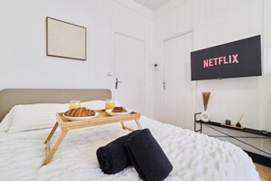 Quarto de hotel com uma cama, um suporte com croissants e suco, uma tela de TV exibindo 'NETFLIX' e uma moldura com um quadro na parede.