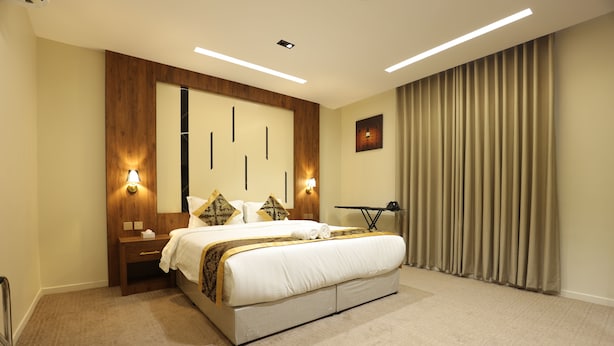 Habitación de hotel con una cama grande, lámparas de noche, un cabecero de madera, un espejo amplio, un escritorio con lámpara y vistas al exterior a través de una ventana con cortinas.
