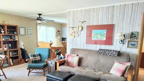 Ein gemütliches Wohnzimmer mit einer Couch, Hocker, einem Sessel, einem Bücherregal, einer Lampe und einem Deckenventilator.