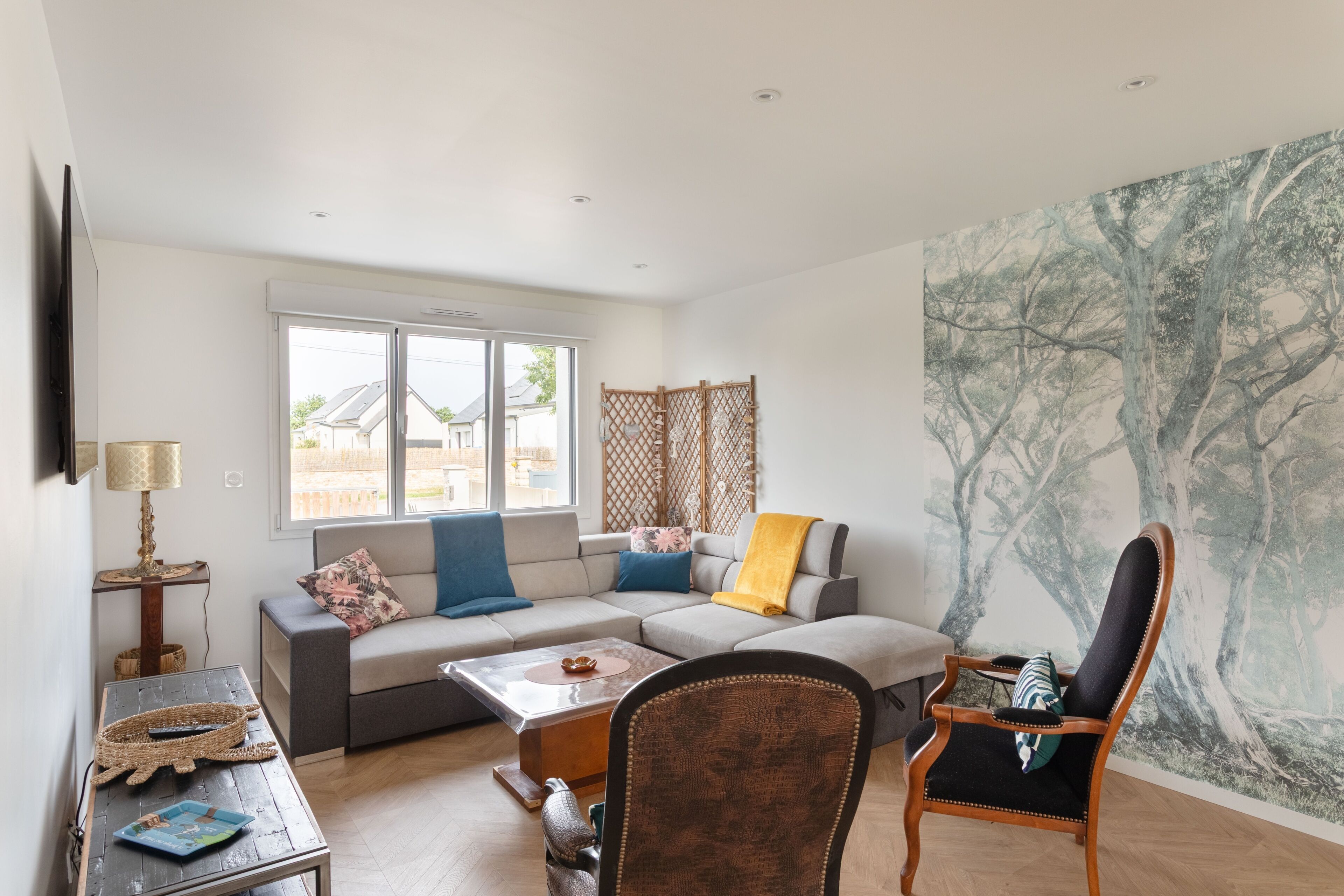 A living room with a sofa, coffee table, and a chair. There is a window with a view of houses outside.
