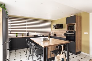 A modern kitchen with a wooden island, black bar stools, a built-in oven, and a large window with blinds. - La Brise - À moins d’1 km de la plage (Fréhel)