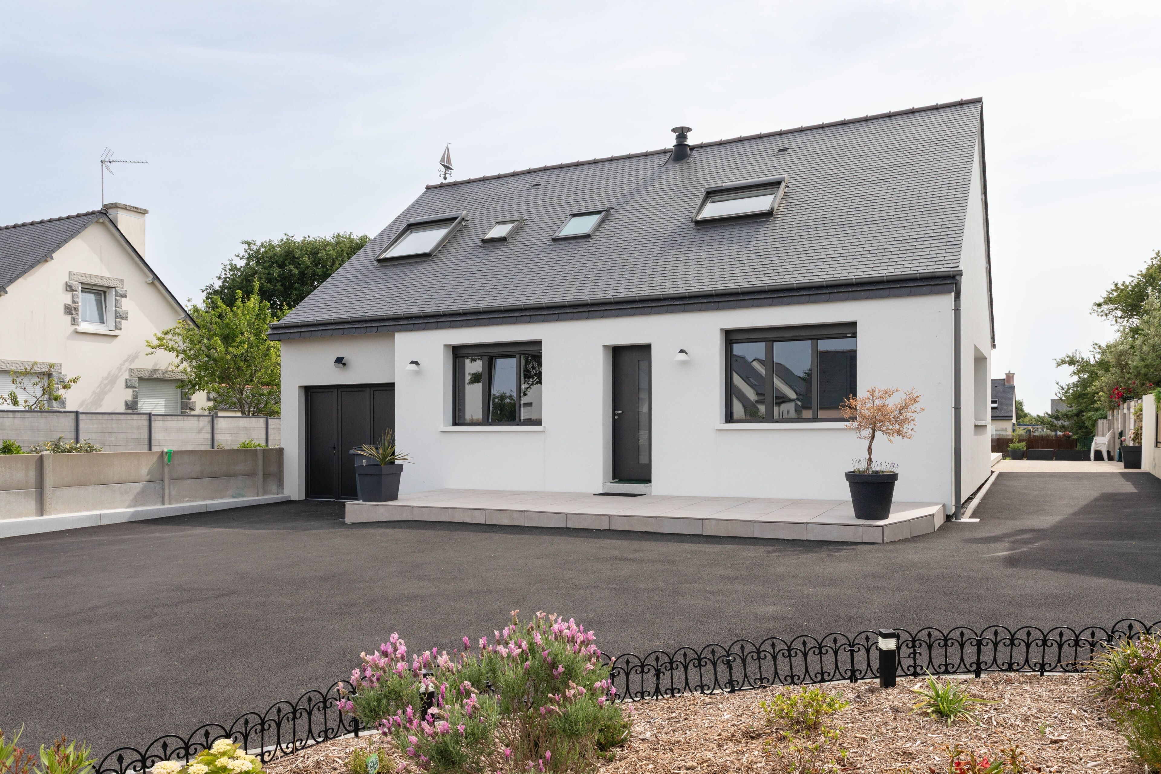 Une maison moderne à deux étages, dotée d’un toit en pente, de grandes fenêtres et d’une clôture en métal noir en premier plan.