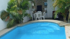 Una piscina con borde de azulejos, rodeada de sillas y una mesa blancos, con palmeras y un muro de piedra al fondo.