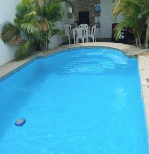 A swimming pool with a tiled edge, surrounded by white chairs and a table, with palm trees and a stone wall in the background. - La Casa Ideal para tus Vacaciones en Playas. Somos tu casa alojamiento temporal. (general villamil playas)