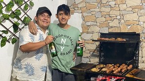 Dos personas junto a un muro de piedra con una parrilla, sosteniendo botellas de cerveza.
