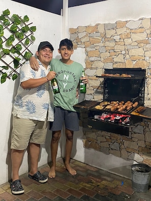 Two people standing next to a stone wall with a grill, holding beer bottles. - La Casa Ideal para tus Vacaciones en Playas. Somos tu casa alojamiento temporal. (general villamil playas)