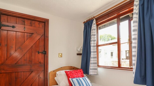 Ein Zimmer mit Holztür, einem Bett mit weißer Bettwäsche und zwei Kissen, einem Fenster mit blauen Vorhängen und Blick auf einen Innenhof.