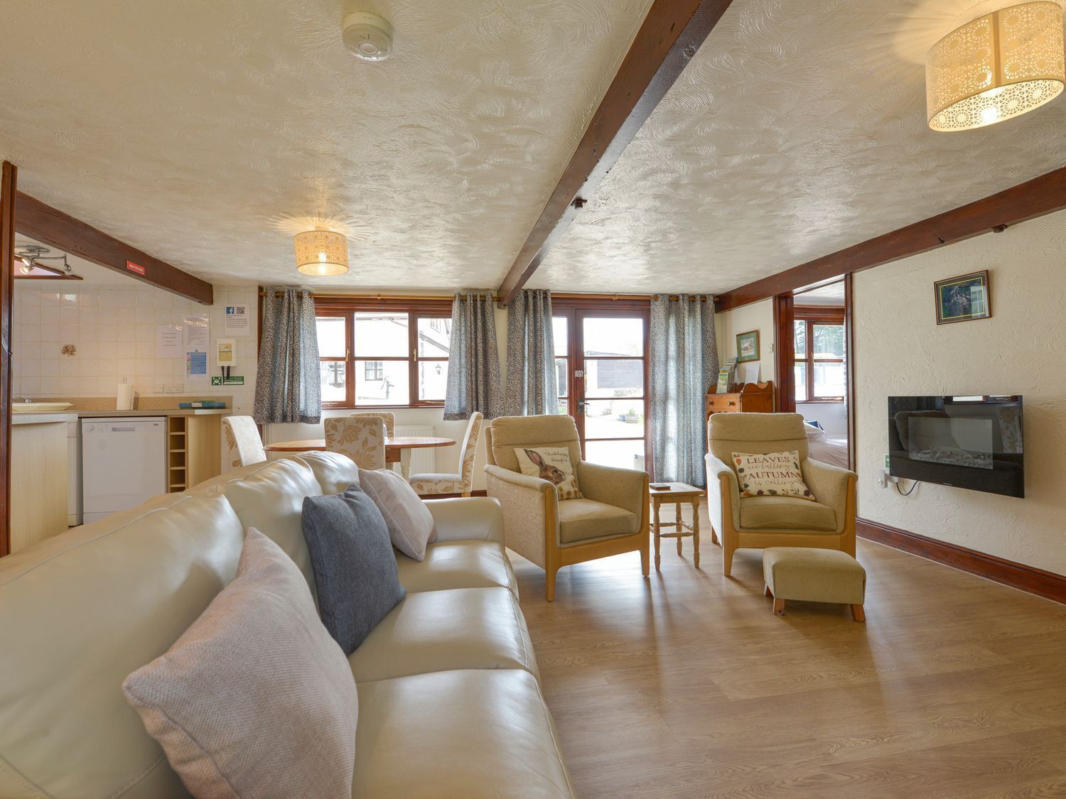 A living room with a sofa, armchairs, a small table, and a TV. There is a kitchen area with a refrigerator and a window.