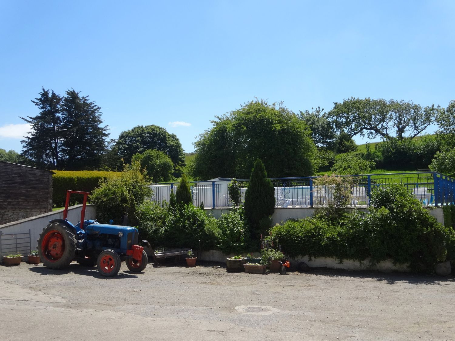 Un trattore blu parcheggiato su un’area sterrata, con un edificio e una zona recintata con vegetazione sullo sfondo.