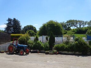 A blue tractor parked in a gravel area with a building and a fenced area with greenery in the background. - Pillhead Cart Linhay (Bideford)