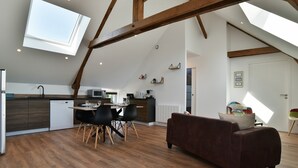 Un espace de vie de style « loft », avec des poutres apparentes, un coin repas comprenant une table et des chaises, une cuisine équipée d’appareils modernes et une grande verrière.