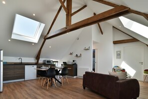 Ein Loft-Zimmer mit freiliegenden Holzbalken, einem Essbereich mit Tisch und Stühlen, einer Küche mit modernen Geräten und einem großen Dachfenster.