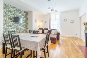 A dining area with a table and chairs, a sofa, a TV mounted on a wall, and a window with curtains. - La Motte Forte - Jolie maison en centre médiéval (Arnay-le-Duc)