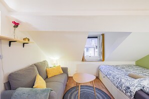 Ein gemütlicher Raum mit Schlafgelegenheit, Sofa, Tisch, Fenster und Heizkörper.
