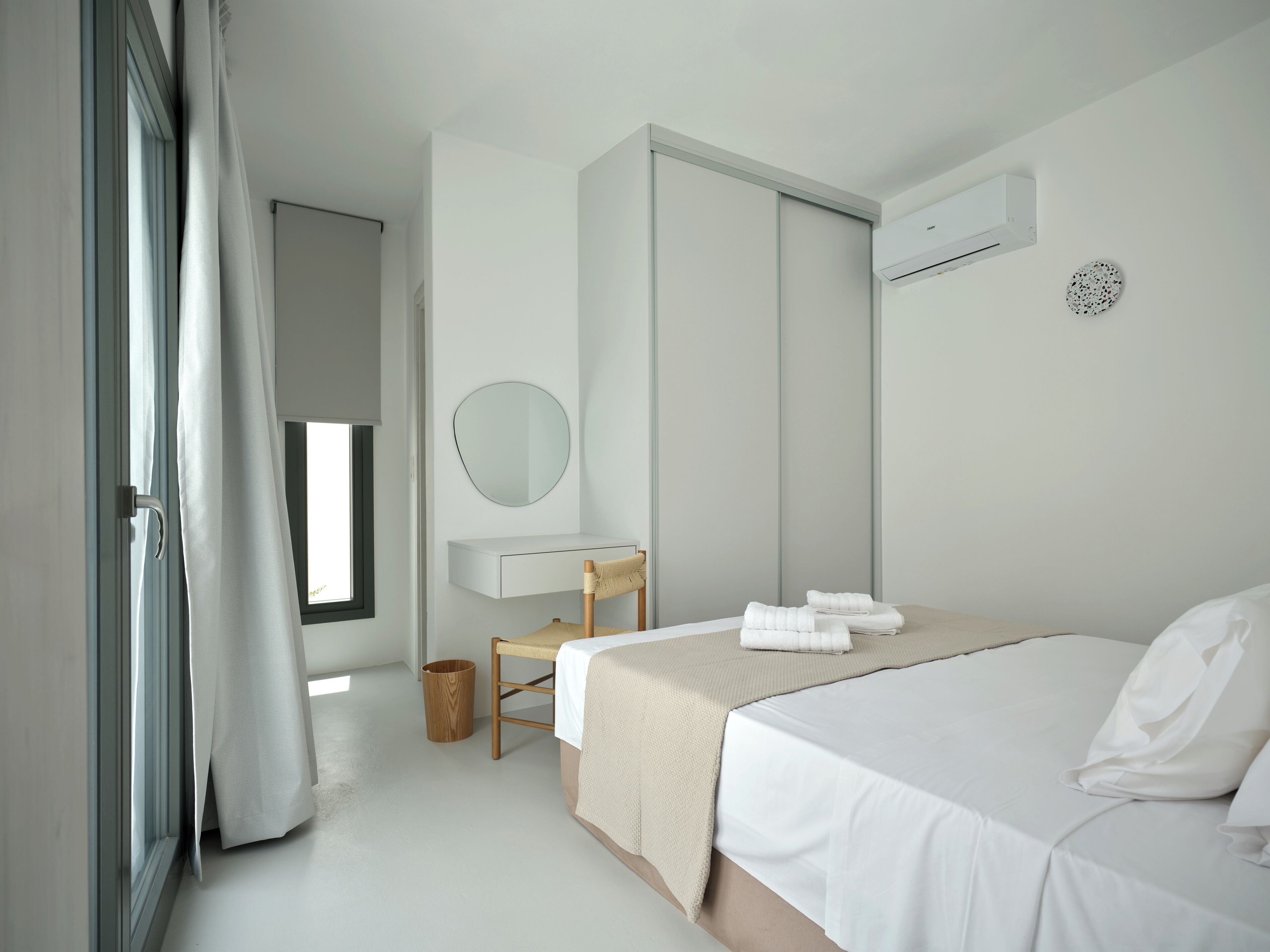 A bedroom with a bed, a bedside table, a chair, a mirror, a window, and an air conditioning unit.