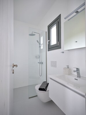 A modern bathroom with a glass shower enclosure, a toilet, a sink with a mirror, and a small window. - Villa Iliada - Paros Escape (Paros)