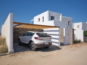 Une voiture blanche est garée sous une pergola en bois devant un bâtiment blanc aux murs et à la porte en pierre.