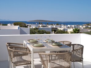 A terrace with a table set for dining, wicker chairs, and a view of the sea and buildings. - Villa Iliada - Paros Escape (Paros)