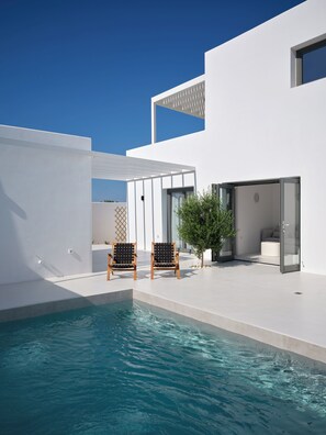 A modern pool area with two wooden chairs, a glass door leading to a room, and a clear blue sky. - Villa Iliada - Paros Escape (Paros)