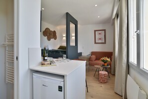 A compact living space with a kitchenette, a dining area, a sofa, and a window with curtains. - Le Palais des Arcades - Studio 100m de la Plage (Fréhel)