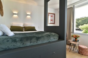 A modern bedroom with a bed, two wall-mounted lights, a framed picture, a small table with a vase of flowers, and a window with curtains. - Le Palais des Arcades - Studio 100m de la Plage (Fréhel)