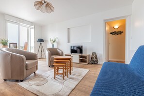 A modern living room with a sofa, chairs, a TV, and a door leading to another room. - Les Voiles de Nacre - Appt à 100m de la plage (Courseulles-sur-Mer)