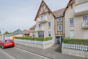 A residential building with a balcony, a gated driveway, and a red car parked on the street. - Les Voiles de Nacre - Appt à 100m de la plage (Courseulles-sur-Mer)