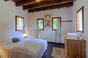 A bedroom with a bed, bedside table, lamp, wooden cabinet with a sink, and a window with curtains. - La Parenthèse Fleurie - Avec Piscine Privée (Saint-Priest-les-Fougères)
