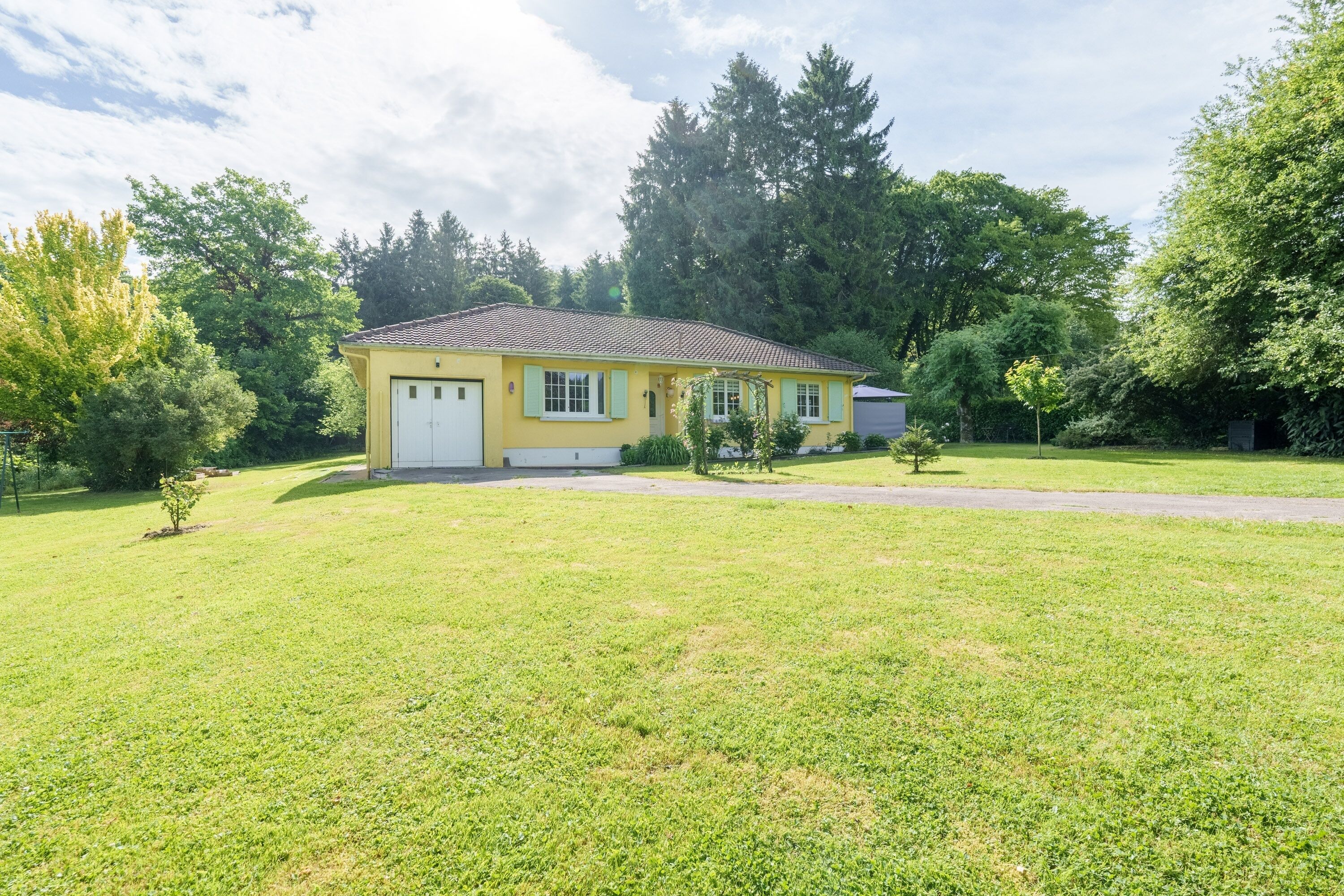A single-story yellow house with a white garage door, surrounded by a well-maintained lawn and trees.