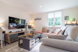 A modern living room with a flat-screen TV, a sofa, a coffee table, and a window with a view of greenery.