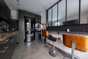A modern kitchen with dark cabinetry, a black tiled backsplash, and two bar stools with wooden seats. - La Villa Rosa - Avec Piscine Privée (Toulouse)