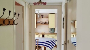 Un couloir mène à une salle à manger où l’on trouve une table recouverte d’une nappe à rayures, un luminaire suspendu au plafond et un crochet fixé au mur.